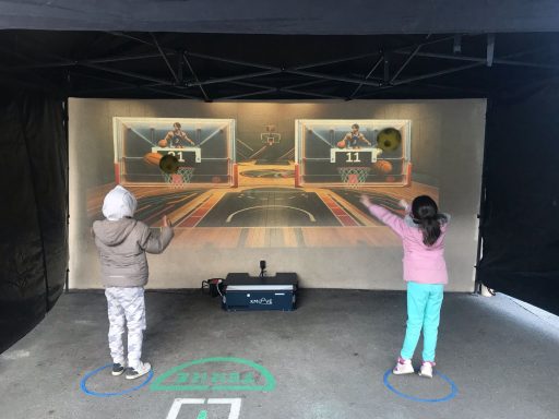 Deux enfants interagissent avec un jeu de basketball sur un mur, en imitant des tirs.